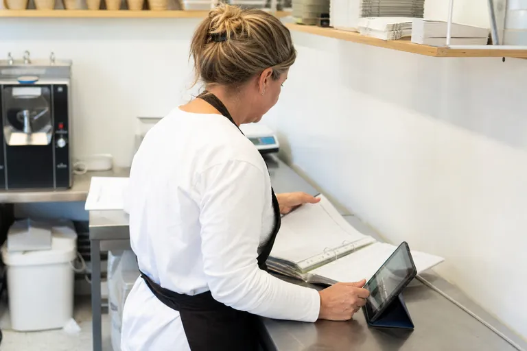 Inhaberin vergleicht Papierlisten in einem Ordner mit digitaler Dokumentation auf einem Tablet